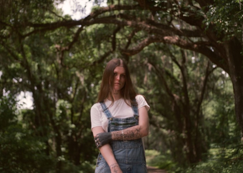 A person with long brown hair wearing a white tshirt and blue denim overalls in a woodsx